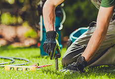 Person mit Handschuhen arbeitet im Garten und steckt einen Pflanzstab in den Boden.