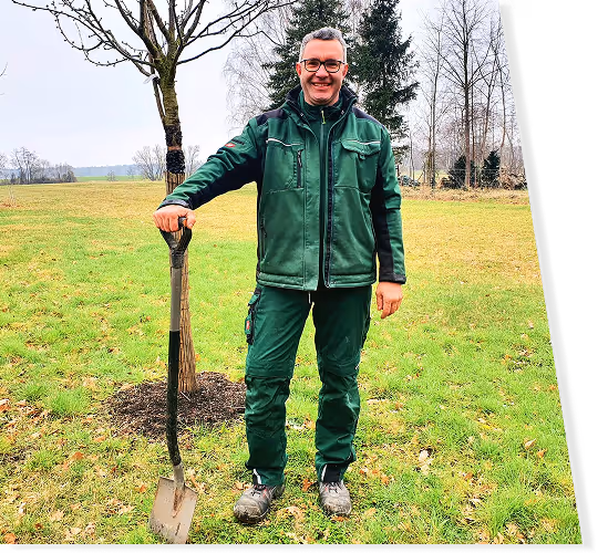 Mann in grüner Arbeitskleidung steht auf einer Wiese und lehnt sich an eine Schaufel.